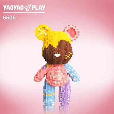 A Brick Bear toy with yellow head, white speckles, pink ears, and floral-patterned blue/purple limbs on a gradient pink backdrop.